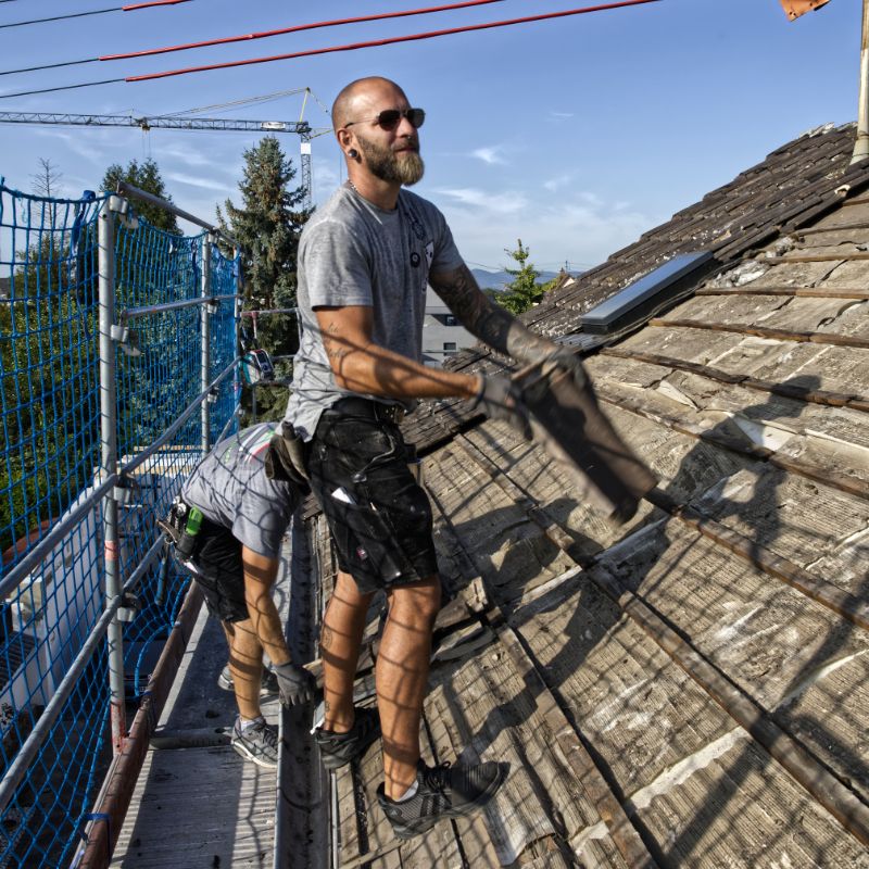 Dachdecker mit Sonnenbrille bei der Arbeit auf einem Dach, dahinter ein Gerüst